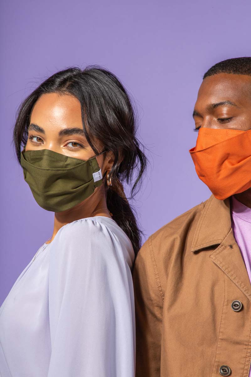 Two people stand side by side against a purple background, both wearing face masks—one in olive green and the other in orange. The person on the left wears a light blouse; the one on the right wears a brown jacket.