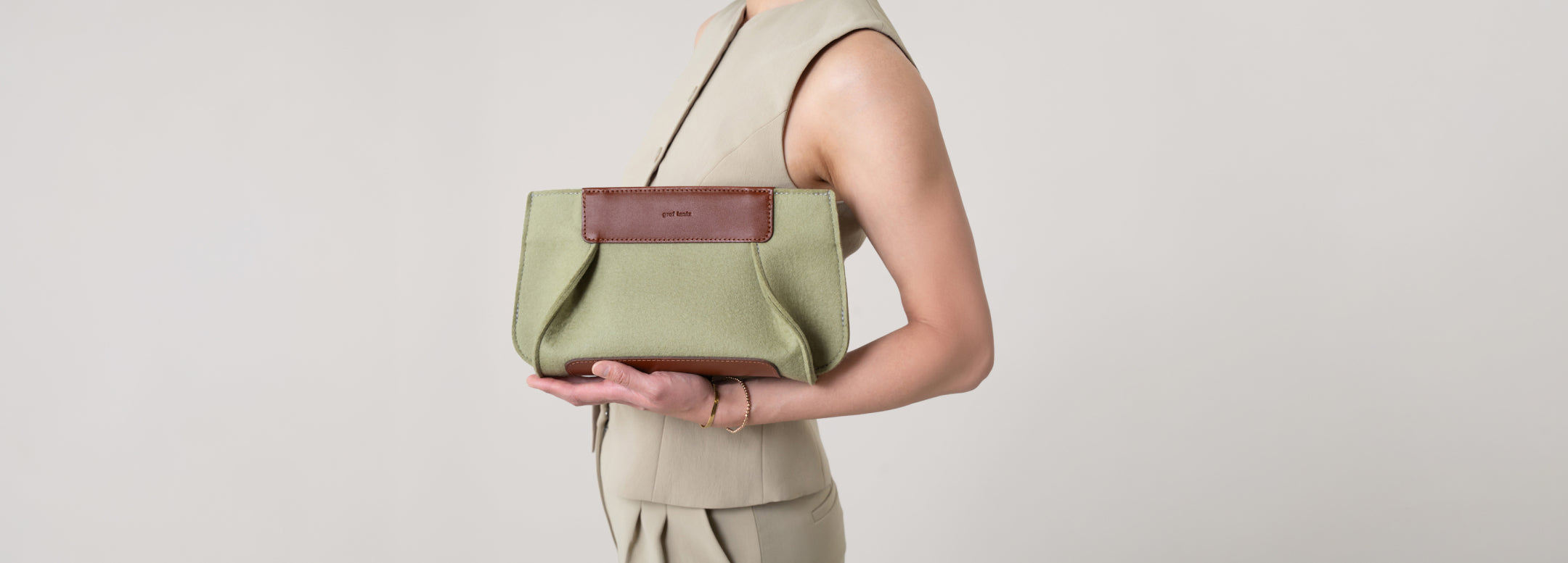 A woman in an off-the-shoulder cream dress sits on a rock against a light wall, placing a cellphone into a tan handbag. A leafy branch casts a shadow nearby.
