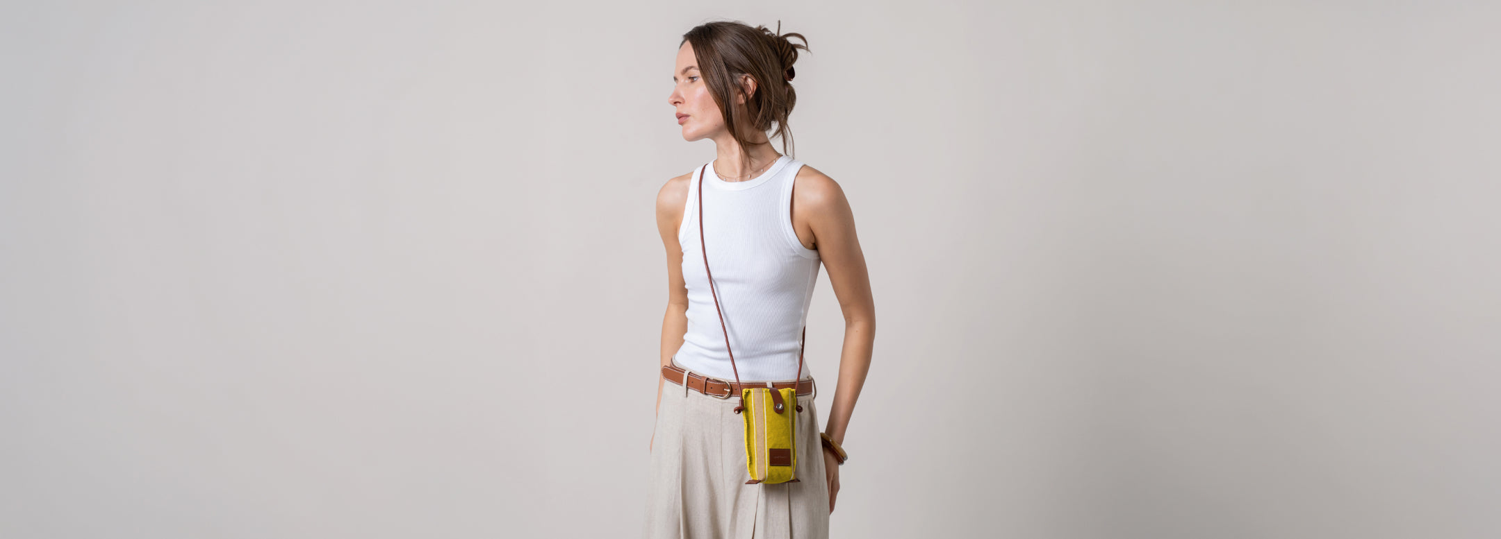 A person in a light beige outfit holding a brown crossbody bag with a beige flap, adjusting the strap. The surroundings are a neutral, soft background.