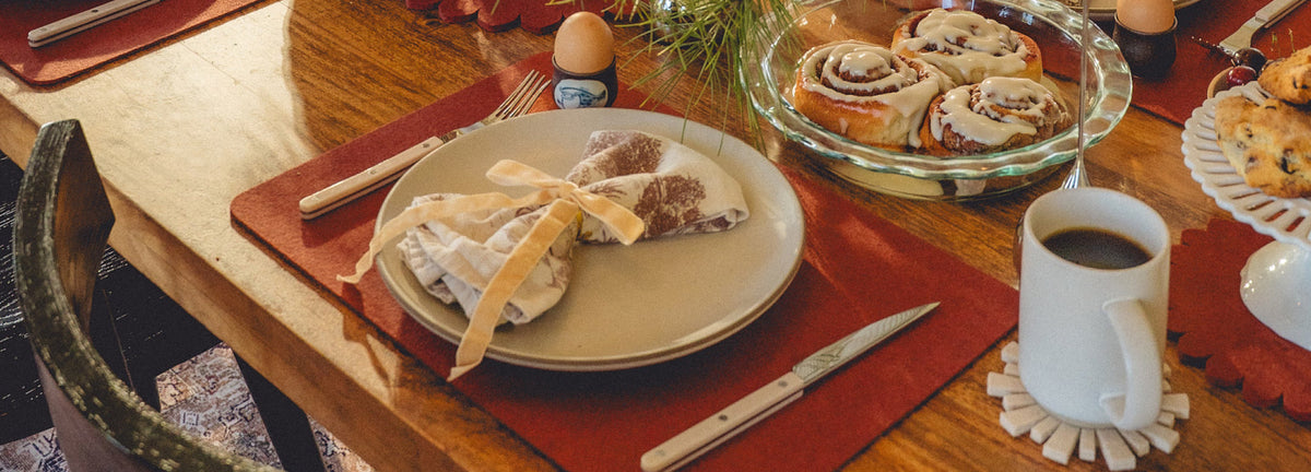 A place setting at a breakfast table featuring a cloth-wrapped napkin, a glass dish of cinnamon rolls, a boiled egg, and a mug of coffee.