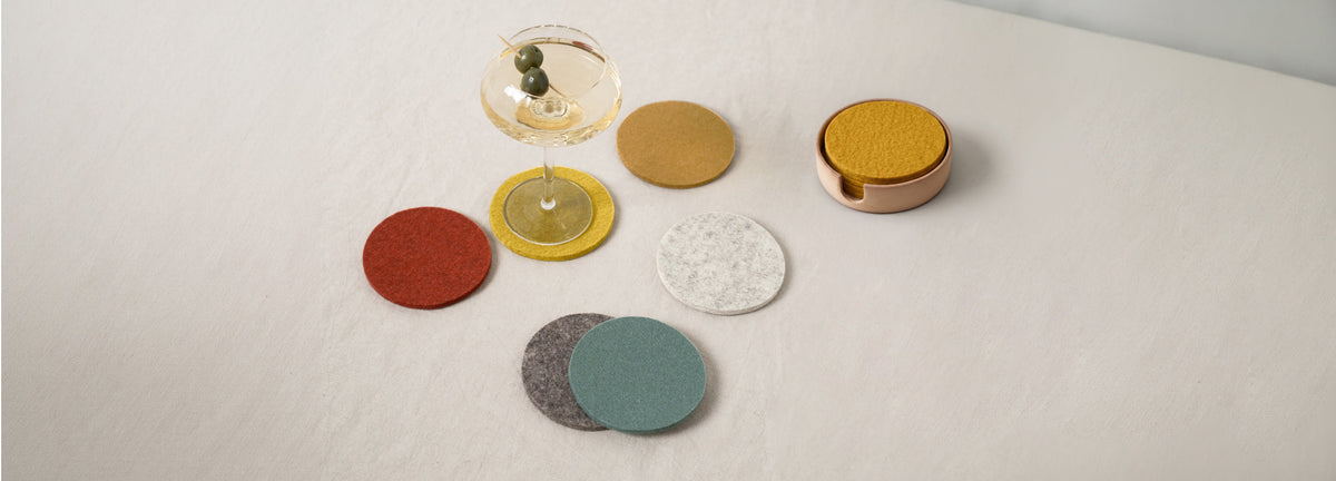 A martini glass with olives sits on a yellow felt coaster, surrounded by five colorful coasters and a copper container holding more coasters on a light surface.