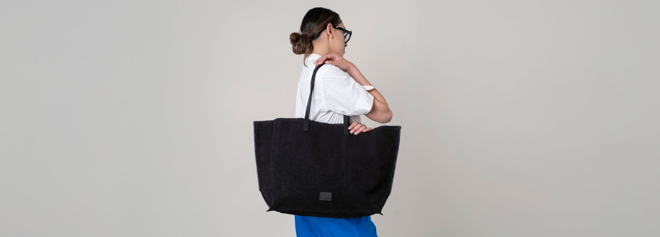 A person carrying a large, structured black tote bag with black handles. The person is wearing black pants and heeled sandals, walking against a sunlit concrete wall.