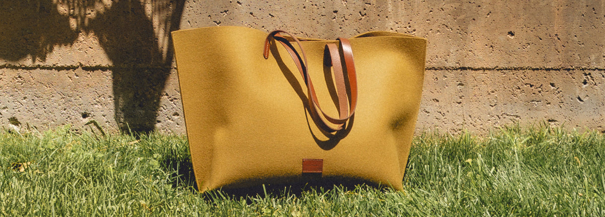 A large tan tote bag with brown handles sits on green grass in front of a concrete wall under sunlight.