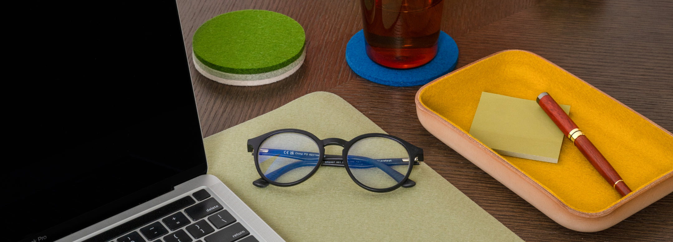 A laptop with a partially visible Apple logo sits on a scalloped mat on a wooden table. Nearby are a closed laptop sleeve and two pouches stacked on books. Soft shadows from tree branches dapple the scene.