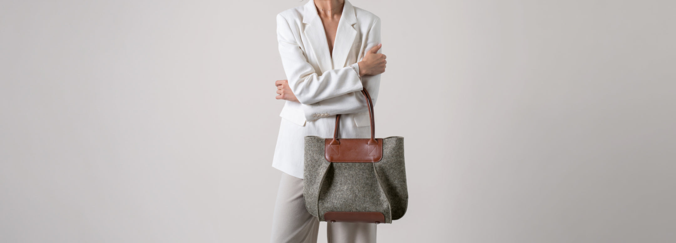 A person wearing a cream sweater and beige pants holds a large olive-green tote bag with tan leather handles and accents against a plain light background.
