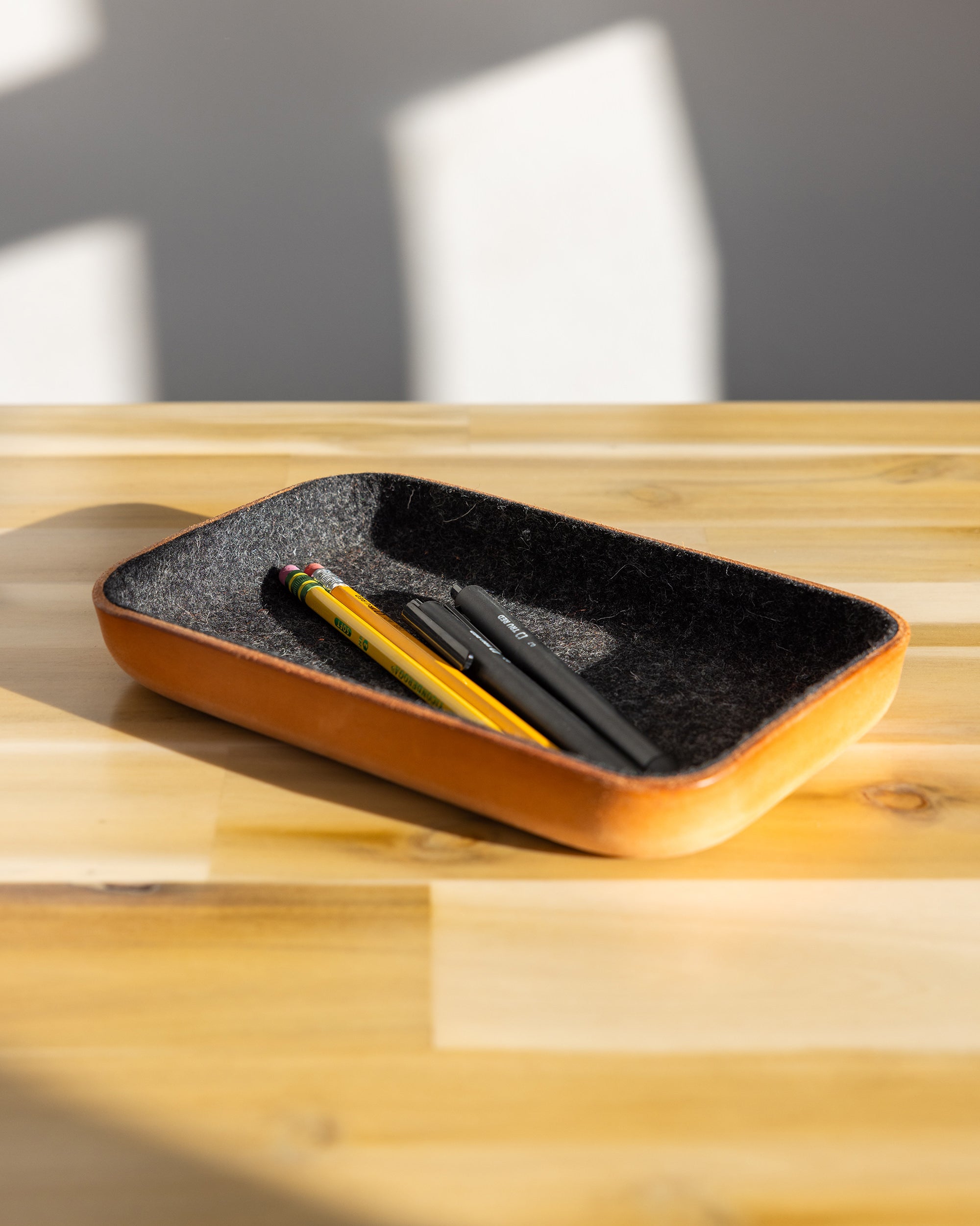 A graf lantz rectangular Vachetta leather catchall tray with a Merino wool interior sits on a wooden surface. Inside are two pencils, one yellow with an eraser and one black. Sunlight casts soft shadows, creating a warm and serene atmosphere.