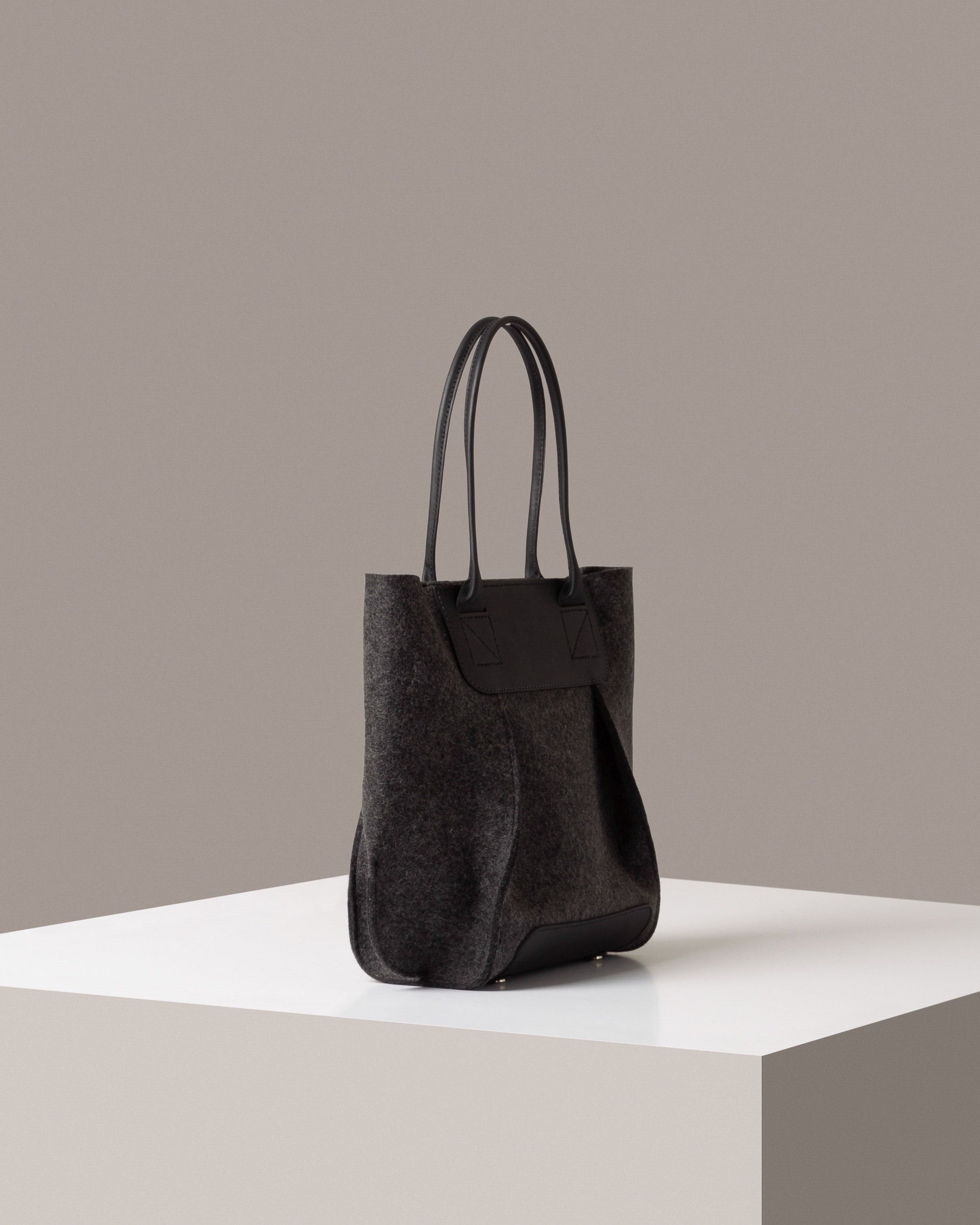 A dark grey Frankie Merino Wool Felt Midi mid-sized tote bag on a white base displayed in a side-view
