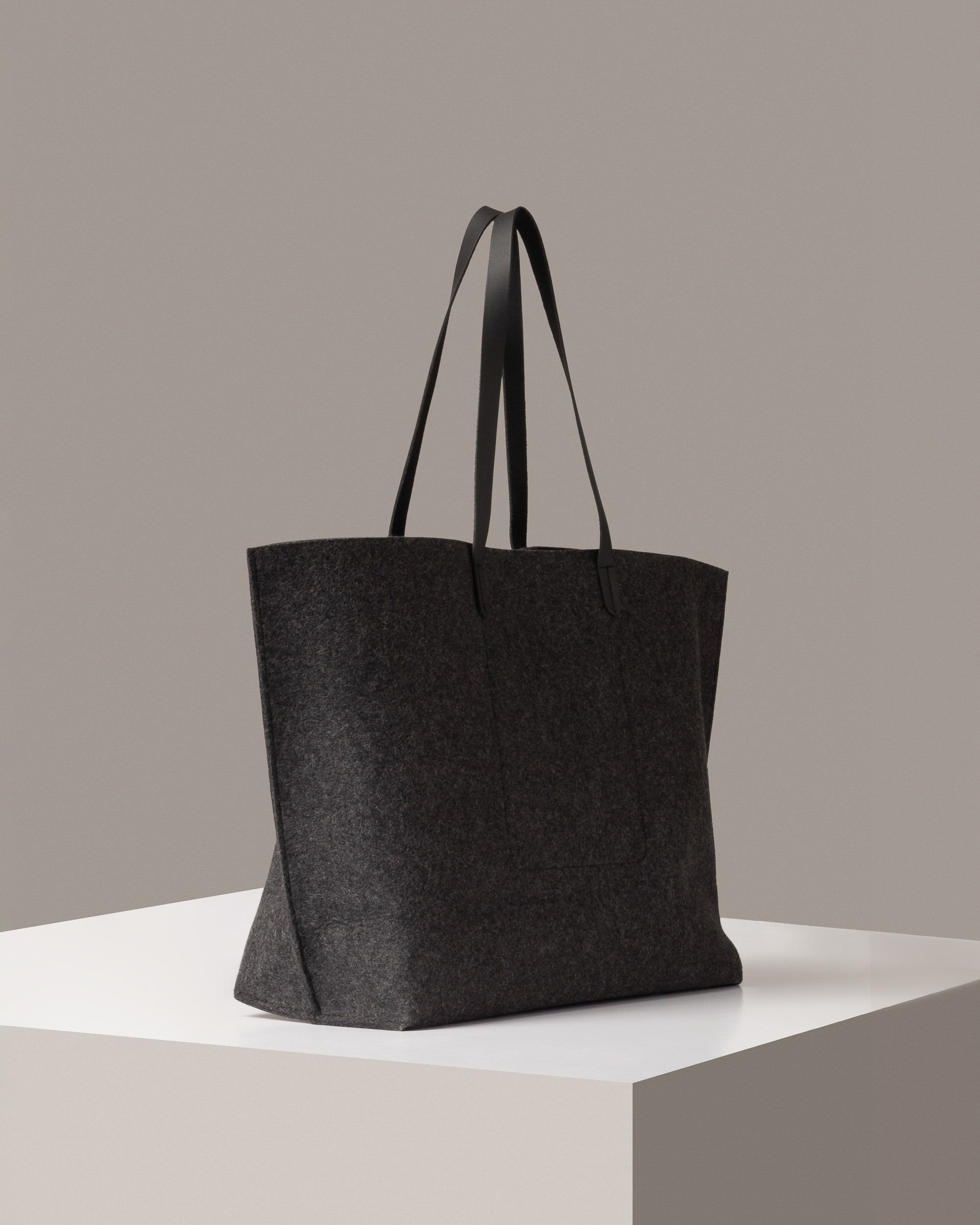 A dark grey Hana Merino Wool Felt Boat Bag on a white base displayed in a side-view