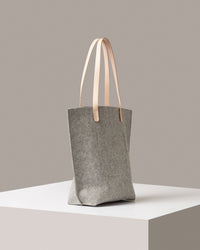 A grey felt tote bag with beige leather handles on a white base displayed in a side-view