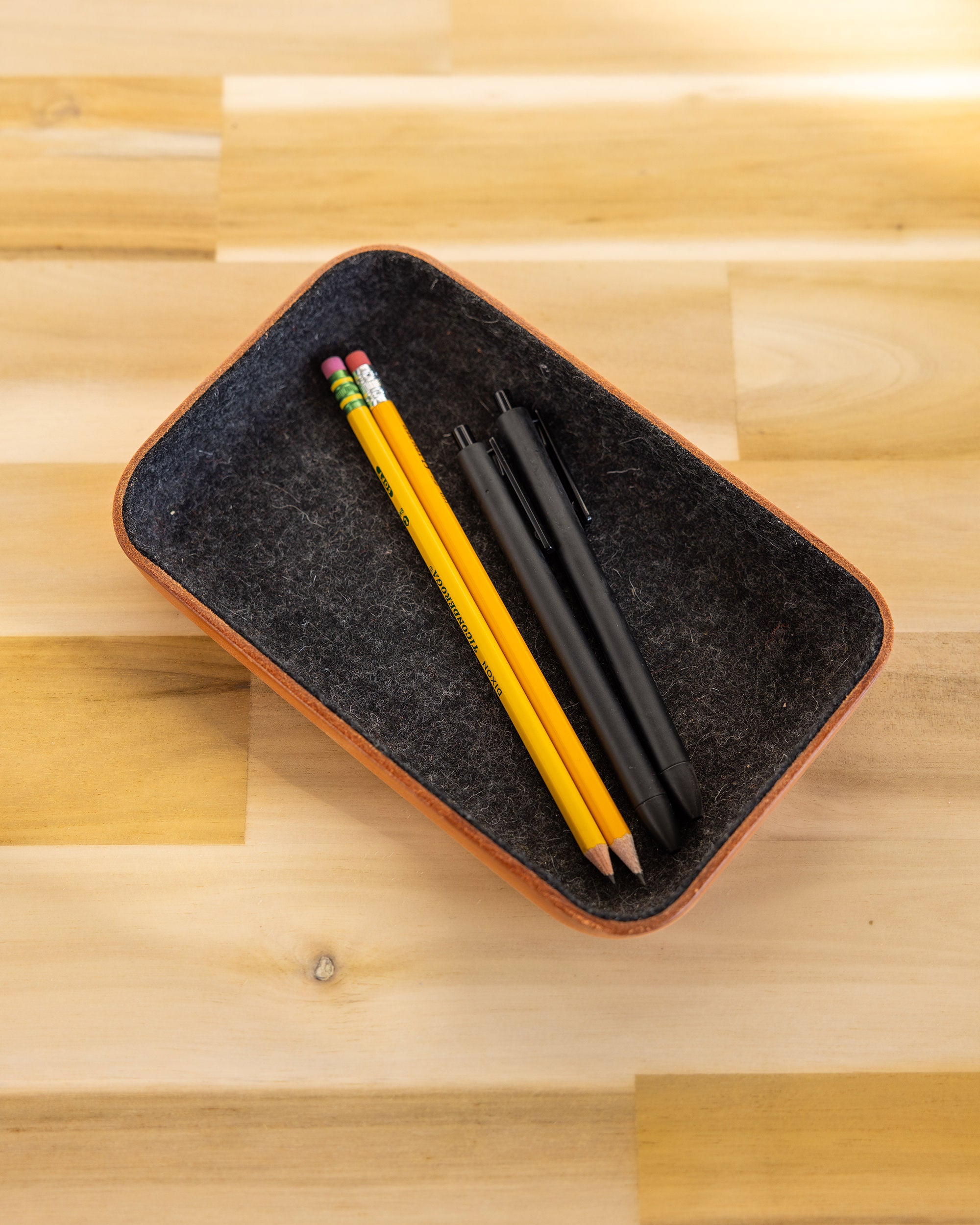 A graf lantz Vachetta leather and felt catchall tray rests stylishly on a wooden surface, holding two yellow pencils with pink erasers and two black pens.