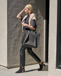 A woman carries a charcoal-colored Frankie Merino Wool Felt tote bag over her arm while walking, showcasing its chic design