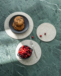 A plate of cookies and a bowl of red berries rest on graf lantz gray Merino wool mats placed on a green marble surface. A fork and scattered berries complete the arrangement, perfectly accentuating the heat-resistant trivet under soft light.
