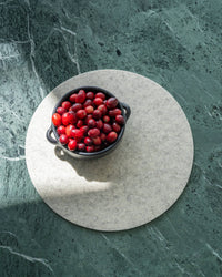 A bowl of red cranberries rests on a round, light-colored graf lantz placemat made from biodegradable materials, placed on a green marble surface with natural light creating delicate shadows.