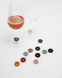 Two wine glasses feature pink liquid being poured, each adorned with colorful Wine-Os markers at the base made from Merino wool felt by graf lantz. Nine more felt rings in different colors are arranged on a white surface nearby.