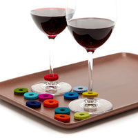 Two glasses of red wine sit on a brown tray, each decorated with vibrant Merino wool felt drink markers by graf lantz. More sustainable luxury markers in various colors are elegantly scattered across the tray.