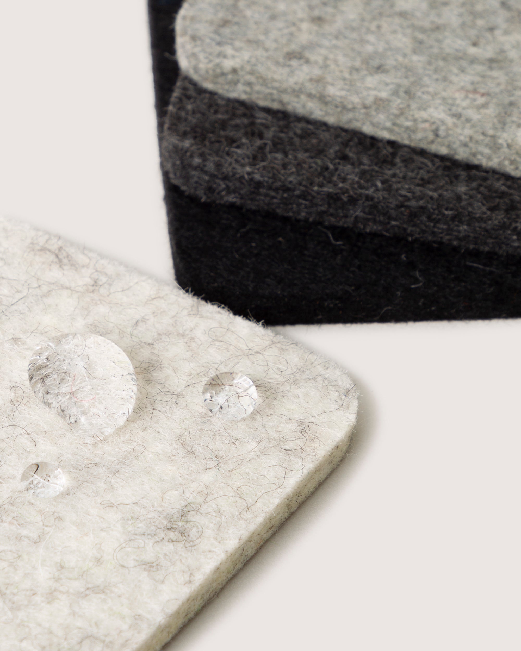 A close-up shows three stacked graf lantz Bierfilzl felt pads, crafted from German Merino wool in light, medium, and dark gray. Drops of water on the top light gray pad emphasize its texture and water-repellent quality against an off-white background.