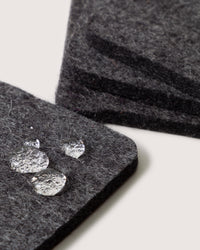 Close-up of four dark gray felt coasters by graf lantz, made from German Merino wool, stacked in the background. Water droplets bead on a foreground coaster, showcasing its water resistance. The scene is set against a neutral backdrop.