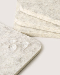 Close-up of three square coasters by graf lantz, crafted from soft Merino wool, stacked on a surface. The foreground coasters water droplets enhance its light gray texture, adding sophistication to any home décor setting.