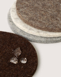 A stack of three graf lantz Merino wool coasters in dark brown, light gray, and dark gray sits against a plain white background. The top brown coaster features several liquid droplets, highlighting the visible texture of the felt as a piece of sustainable home décor.