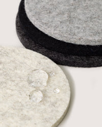 Three round felt coasters, in light gray, dark gray, and black, slightly overlap each other. Made from sustainable and biodegradable German Merino wool by graf lantz, the light gray coaster in front has a few water droplets on it.