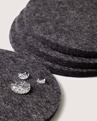 A stack of graf lantz gray felt coasters made from fine German Merino wool showcases three clear, faceted glass objects resembling gemstones on a plain white background.
