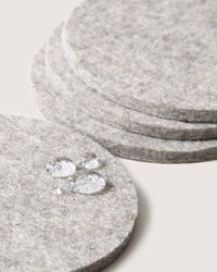 Close-up of a stack of round, gray coasters by graf lantz, crafted from German Merino wool. Water droplets bead on the surface, emphasizing their water-resistant texture against a matching light gray background that enhances their refined appearance.