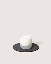 A white candle in a frosted glass holder rests on a round, gray felt Merino wool trivet by graf lantz, offering elegance and heat resistance against the light background, highlighting the simplicity of the setup.