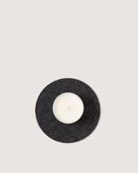 A single white graf lantz candle sits on a heat-resistant trivet against a light gray backdrop. The unused candles wick is perfectly centered, creating a minimalistic and balanced composition.