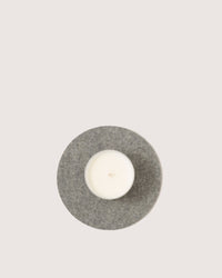 A white candle in a glass holder rests on a round, gray felt mat from graf lantz, made of Merino wool. The minimalist design is enhanced by the mats heat-resistant properties against an off-white background.