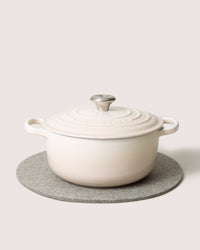 A beige ceramic Dutch oven with a lid sits on a circular gray felt trivet crafted from heat-resistant Merino wool by graf lantz. The pot has two handles and a metallic knob on the lid, all placed against an off-white background.