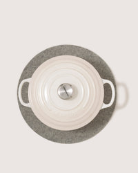 A graf lantz heat-resistant gray felt pad holds a white oval cast iron Dutch oven, viewed from above, featuring two handles and a metal knob on the lid.