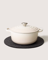 A cream-colored graf lantz cast iron Dutch oven with a lid and heat-resistant metal knob sits on a dark circular trivet, featuring sturdy curved handles and a smooth, glossy finish against a plain light background.
