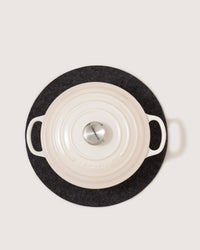 A white cast iron Dutch oven with a silver knob, two handles, and concentric rings on the lid sits on a dark felt trivet from graf lantz that wicks moisture.