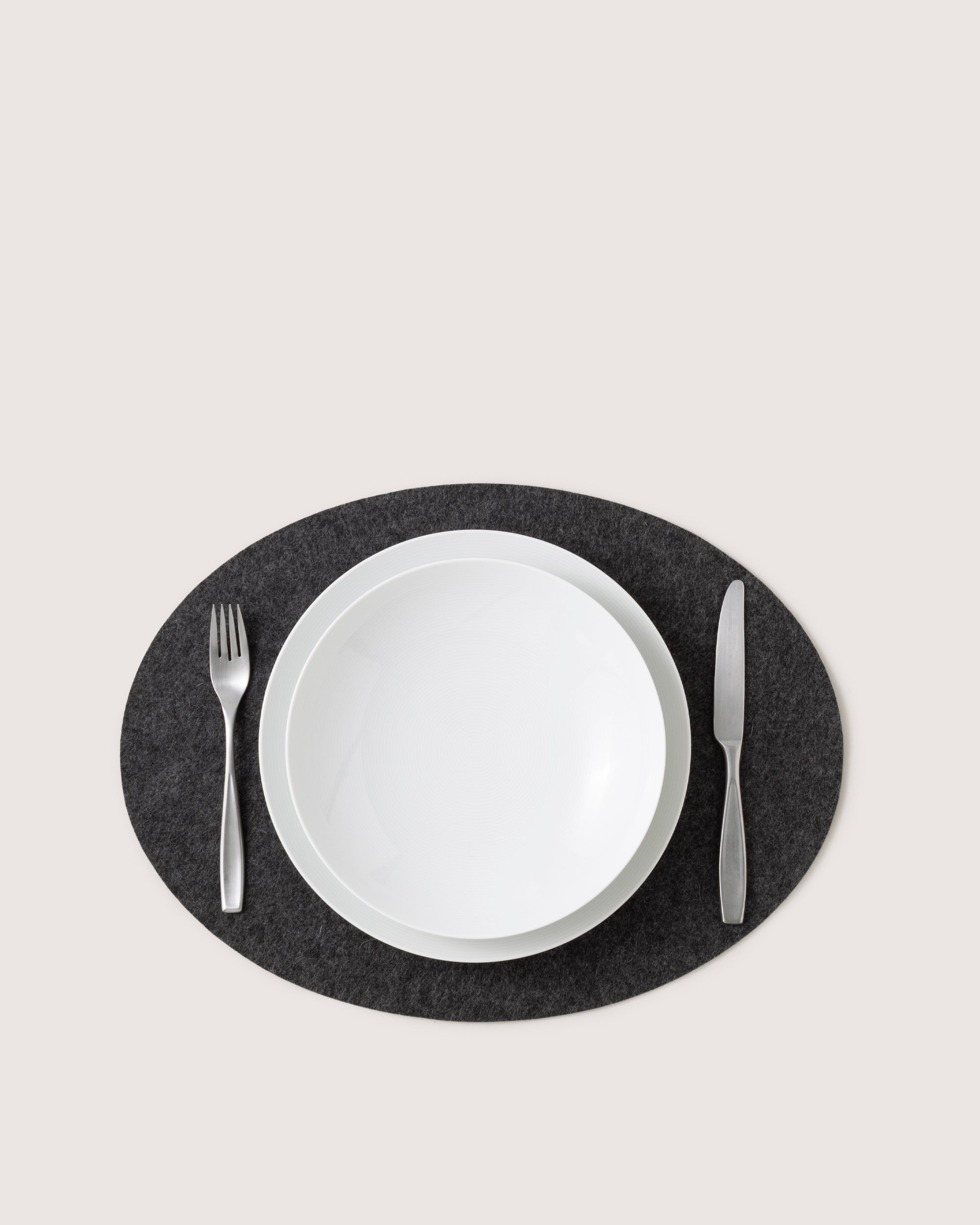 A white ceramic bowl sits on a graf lantz premium Merino wool placemat, contrasting against its dark gray texture. A silver fork is on the left and a silver knife on the right. The light gray background enhances the elegant setting.