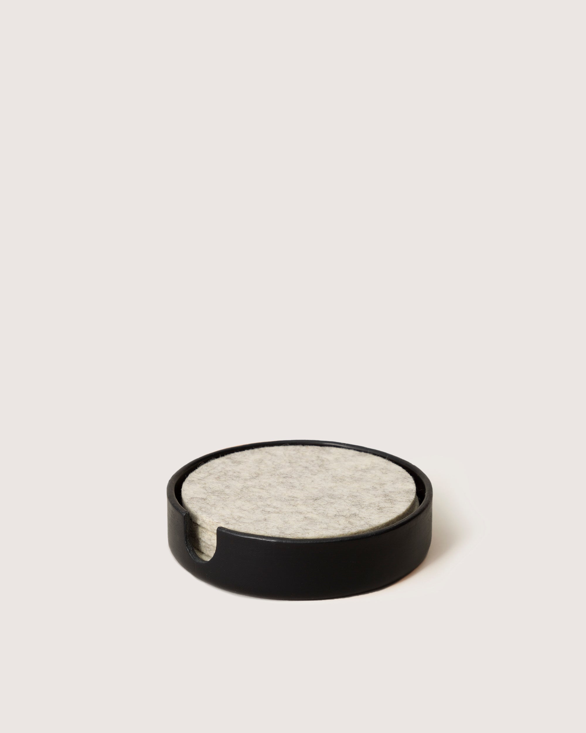 A graf lantz round black sculpted catch-all elegantly holds a stack of circular light gray felt coasters, with one subtly exposed against an off-white background, creating a versatile organizer for any setting.
