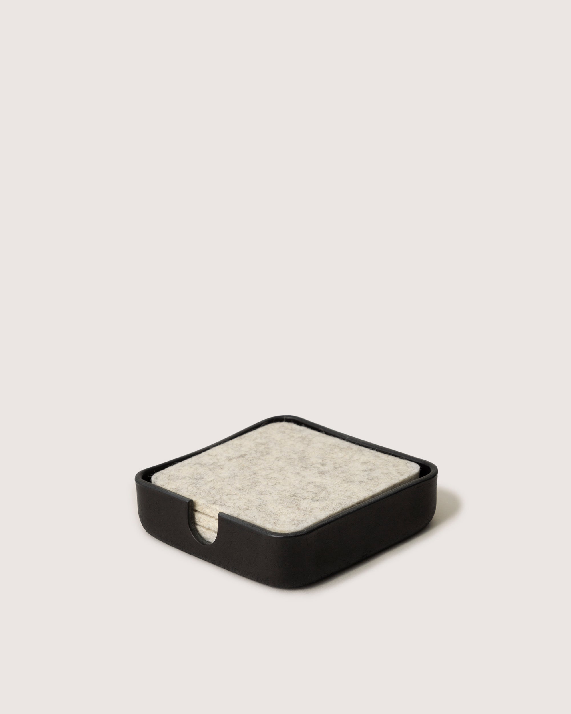 A sophisticated square black coaster holder by graf lantz, crafted with Vachetta leather, elegantly contains a stack of light gray felt coasters on a plain white background.