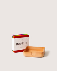 A stack of colorful Merino wool coasters from graf lantz with a Bierfilzl label is displayed alongside a small Kobon leather tray. The coasters, made from sustainable materials, feature vibrant colors that contrast with the neutral tray.