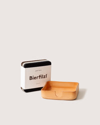 A tan leather tray made from Kobon leather sits beside a stack of square coasters in a white Bierfilzl box. The graphic design by graf lantz features coasters crafted from dark Merino wool, all set against a plain light gray background.