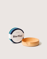 A stack of colorful round felt coasters from graf lantz, made with Merino wool, rests beside a wooden holder featuring a convenient notch. Each coaster is labeled Bierfilzl with a white band, giving an artisanal feel to this handcrafted set from Los Angeles.