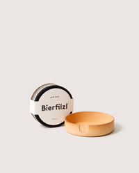 A set of graf lantz circular Merino wool Bierfilzl coasters, wrapped in a white band, rests beside an empty wooden holder. The grayish felt coasters complement the light brown holder with a small side cutout.