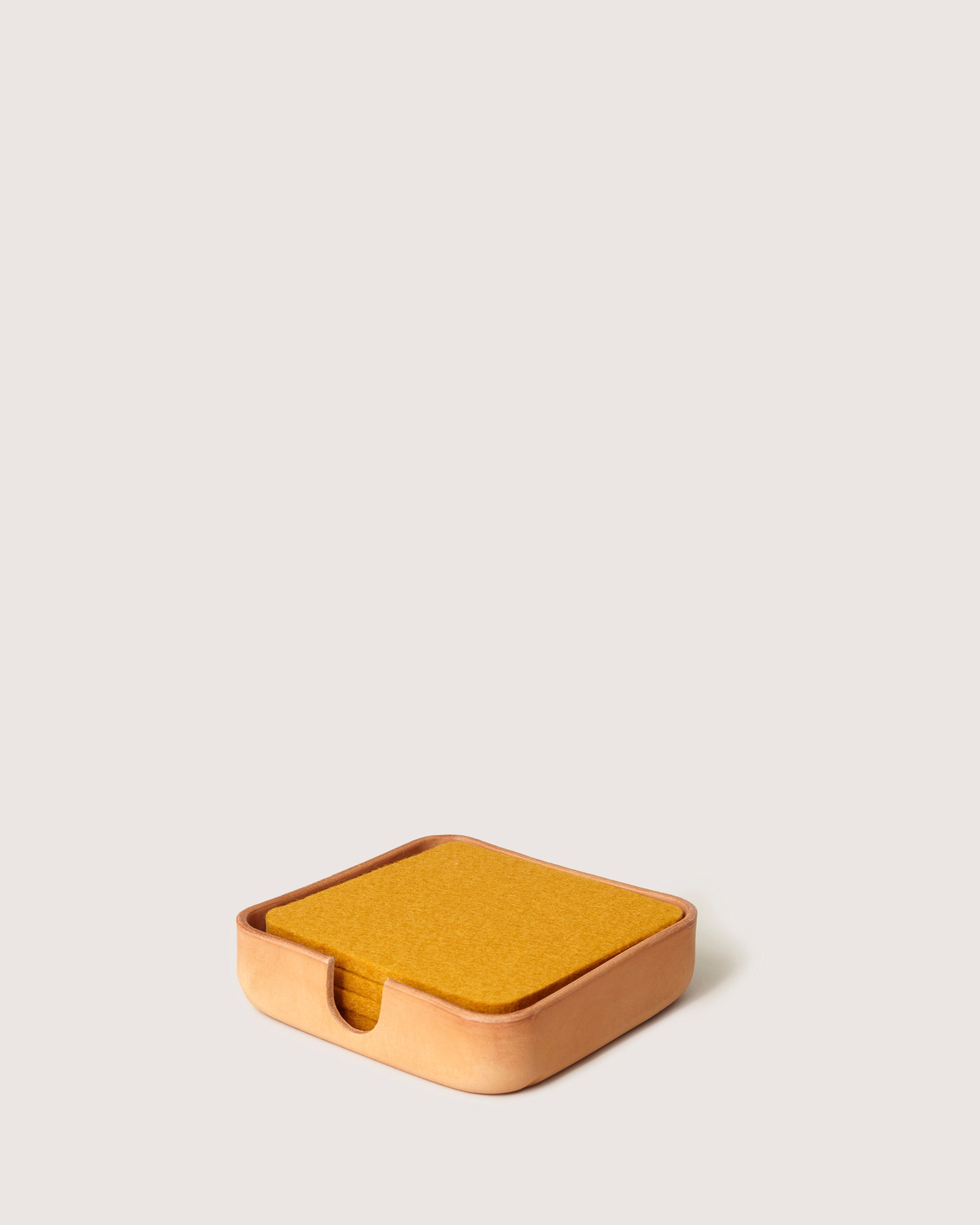 The graf lantz Vachetta leather square coaster holder in tan, set against a light beige background, contains amber felt coasters. Featuring a small notch for easy access, it blends style with functionality effortlessly.