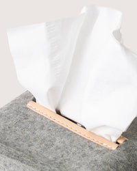 A close-up of a graf lantz Merino Wool tissue box cover showcases a tan leather slit with white tissues partly pulled out. Crafted from Oeko-Tex® Certified materials, it elegantly rests against a neutral, light backdrop.