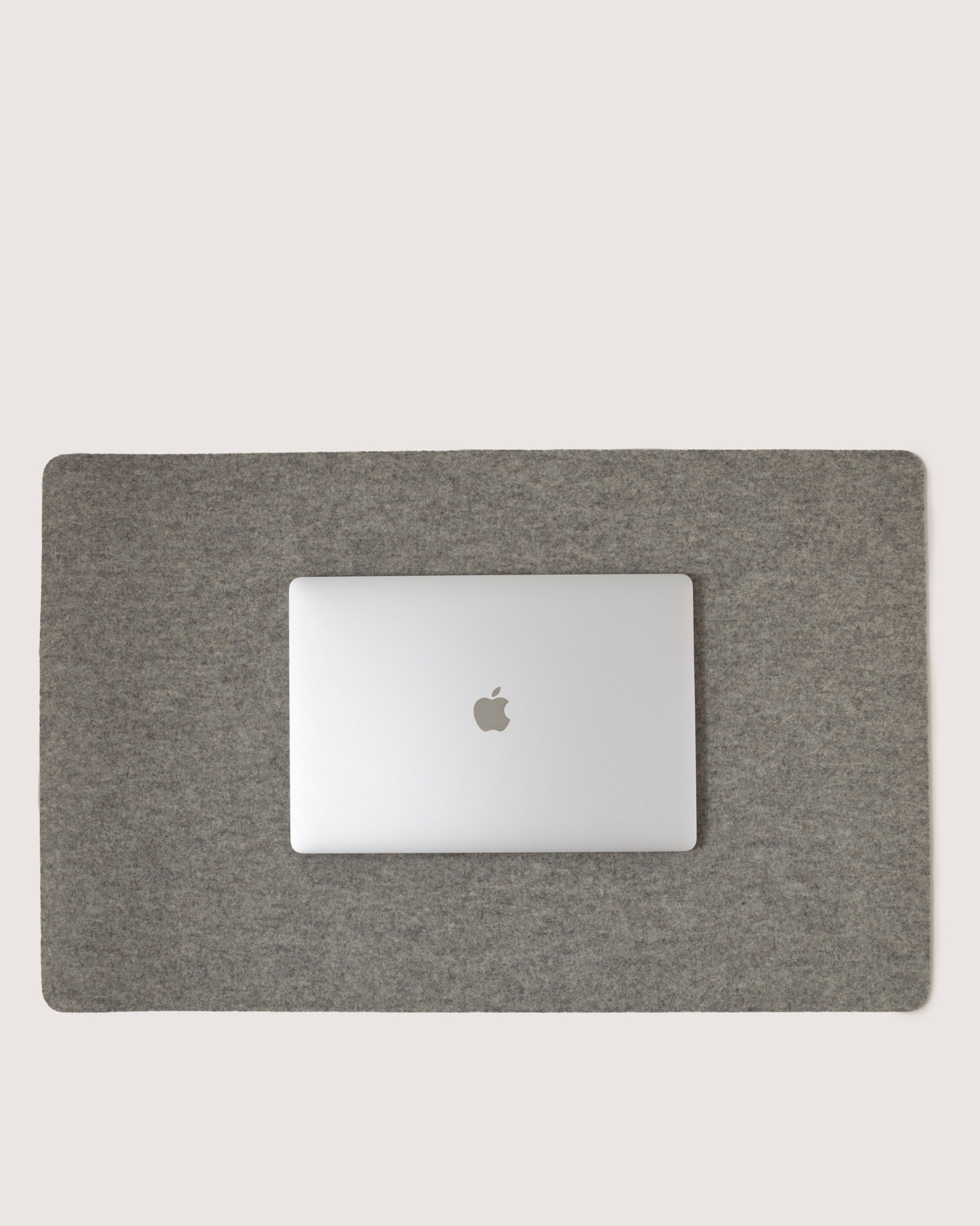 A closed silver laptop with an apple logo rests on a graf lantz Desk Pad crafted from luxurious Merino Wool. The plain background highlights this sleek workspace, emphasizing the elegant contrast between the laptop and the refined gray felt mat.