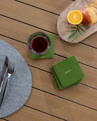 A glass of red liquid rests on a green graf lantz coaster crafted from German Merino wool, alongside a stack of similar coasters on a wooden table. A plate with orange and apple slices sits nearby, while a knife and fork lie on a gray placemat.