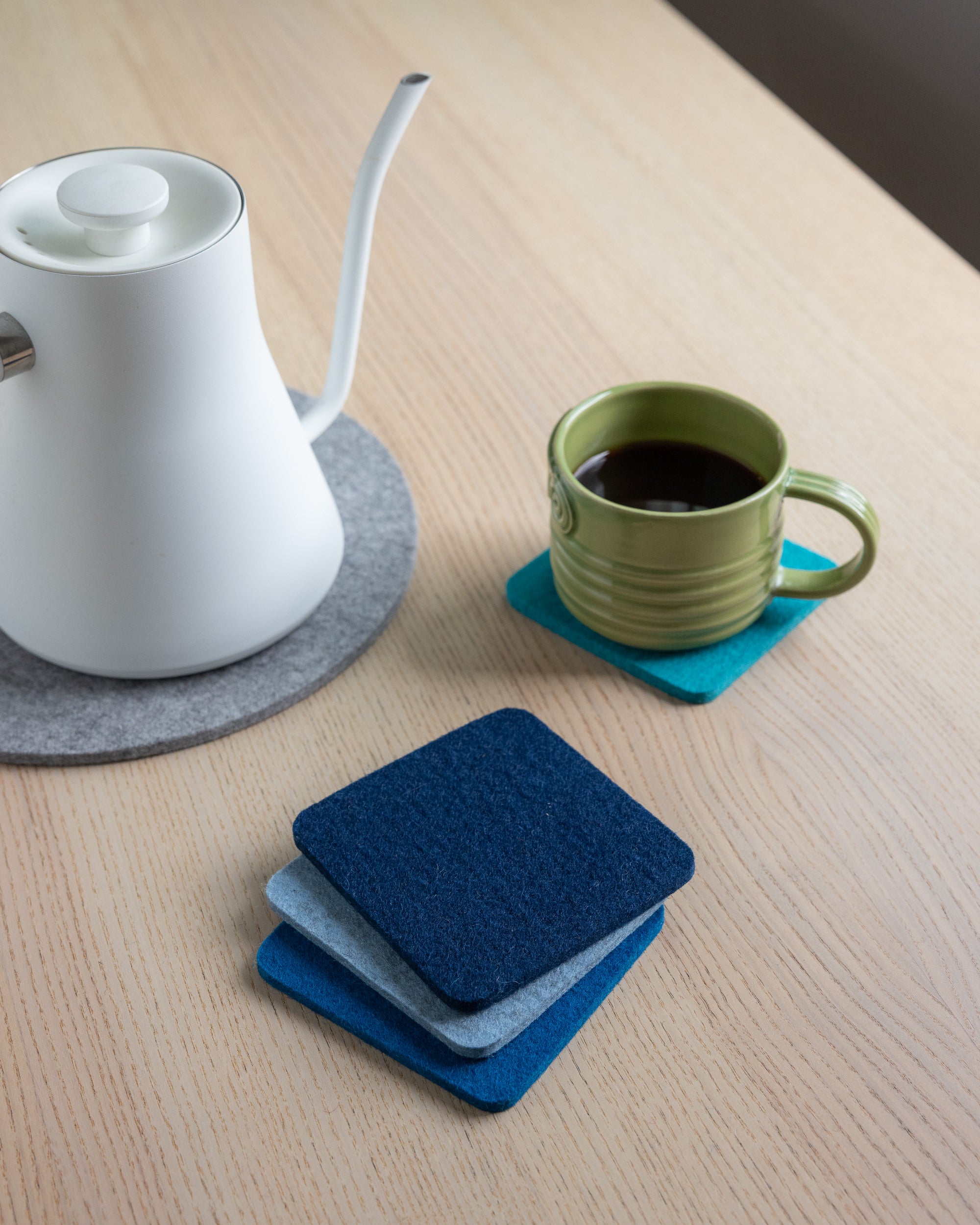A white kettle sits on a gray graf lantz coaster next to a green mug filled with coffee on a blue one. Nearby, three stacked graf lantz Beer Felt coasters in navy, light blue, and teal rest on the light wooden table.