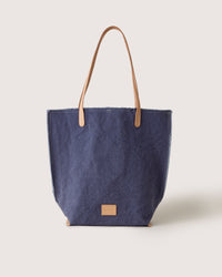 The graf lantz Hana Canvas Tote is a stylish blue denim bag made from durable cotton canvas, featuring tan leather handles and a small tan leather patch on the front, set against a plain light background.