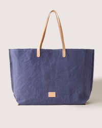Introducing the graf lantz Canvas Boat Bag: a large blue tote with textured fabric and stylish tan leather handles. Its spacious interior provides ample room for essentials, and a small rectangular leather label decorates the bottom center, enhancing its classic design against a light backdrop.