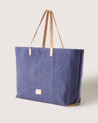 The graf lantz Canvas Boat Bag boasts a stylish blue design with tan leather handles and a beige label at the center. Its elegant exterior opens to reveal a spacious interior, set against a light gray backdrop.
