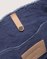 Close-up of a graf lantz blue canvas boat bag with a tan leather strap and metal button. The spacious interior has an embossed leather patch. Slightly frayed fabric edges add to its casual appeal.