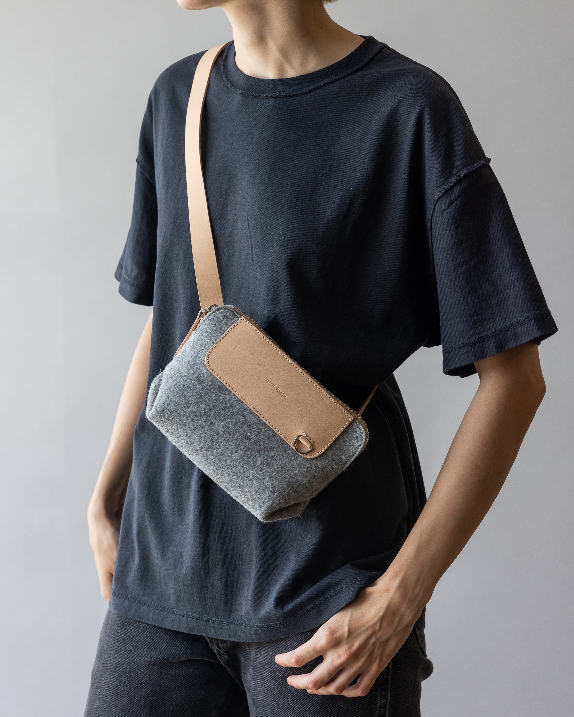 Dressed in a black t-shirt and dark pants, theyre accessorized with a graf lantz beige crossbody belt bag featuring a gray fabric front, hands relaxed by their sides against a neutral gray background for an effortlessly stylish, hands-free look.